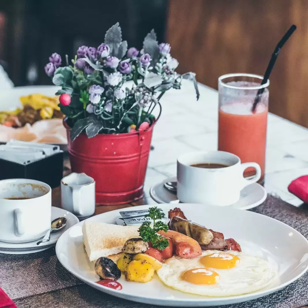 a brunch setting with three cups filled with juice, a bucket pail  with mini roses, two coffee mugs on platters, and a plate of sunny side up eggs, sausage, potatoes and vegetables, cilantro, and toast